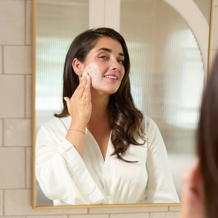 Woman applying cleanser to her face in front of a mirror