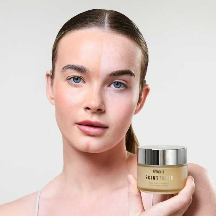 Woman with half of her makeup removed, holding a skincare product with a plain background
