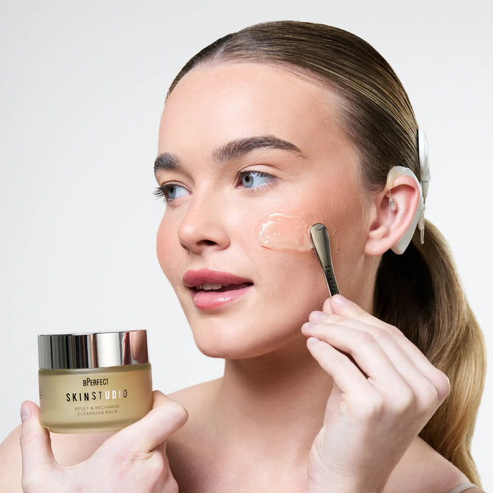 Woman applying cleansing balm with a silver applicator and holding jar labelled 'Skin Studio' on a light background