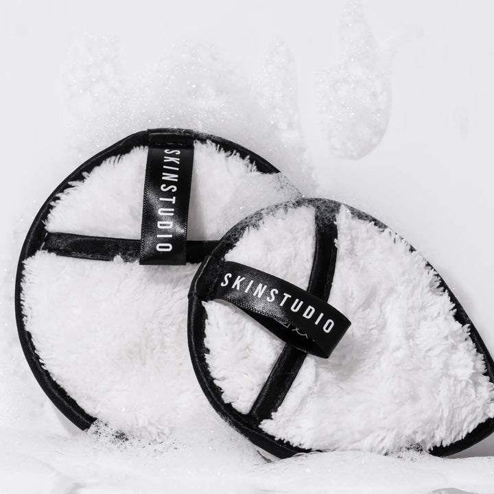 Two round, fluffy cleansing pads with 'SKINSTUDIO' branding on a white background with bubbles.